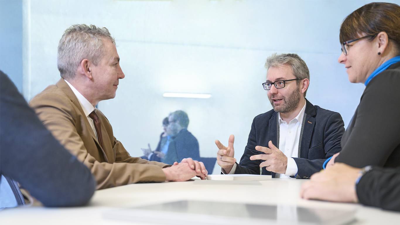 Eine Tarifverhandlung von Paritätischen Arbeitgebern, bei dem drei Personen um einen Tisch sitzen und eine lebhafte Diskussion führen. Das Bild zeigt einen bärtigen Mann in einem braunen Sakko, der gestikulierend spricht, während zwei weitere Teilnehmer zuhören.
